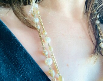 Puede incluir: Un collar largo de cadena dorada con cuentas de perlas blancas. El collar está en capas y tiene un aspecto delicado y femenino.