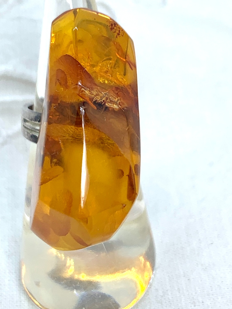 May include: A silver ring with a large, amber gemstone. The amber is a deep, golden color and has inclusions of plant matter.
