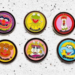 Könnte beinhalten: Sechs runde Pins mit Figuren aus den Muppets. Die Pins sind in einem Kreis auf weißem Hintergrund angeordnet. Die Figuren sind Animal, Kermit, Beaker, Fozzie Bär, Miss Piggy und Gonzo. Jeder Pin hat einen anderen Hintergrund in Farbe und zeigt den Namen der Figur in schwarzer Schrift.