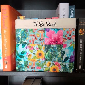 May include: A white fabric storage bin with a blue, green and pink floral pattern. The bin has a beige top with the text "To Be Read" printed in black.