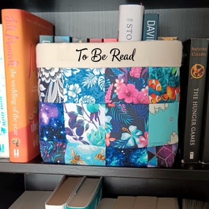 May include: A white fabric storage bin with a patchwork design in shades of blue, pink, and white. The bin has a cream-colored top with the words "To Be Read" written in black cursive.