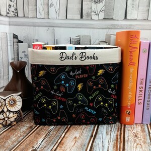 May include: A black fabric storage bin with a white top and the words "Dad's Books" embroidered on the front. The bin is decorated with a repeating pattern of colourful video game controllers and the words "the best player".