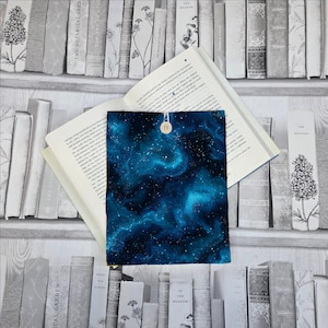 May include: A book sleeve featuring a galaxy design in blue and white, fastened with a wooden button. It's placed over an open book, set against a backdrop of a bookshelf.