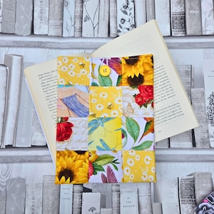 Peut inclure: Une housse de livre patchwork faite à la main, ornée de motifs floraux et naturels. Elle présente des tournesols, des abeilles et des roses rouges, avec une fermeture à bouton jaune. Un livre ouvert est visible.