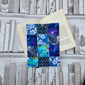 以下が含まれることがあります： ボタン留めのパッチワークブックカバー。花柄、銀河、魚の模様の様々な青色の生地を使用。カバーは開いており、ページが見える本を保持しています。