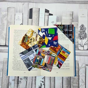 May include: A collection of colorful fabric corner bookmarks with various patterns, including books, text, and abstract designs, are placed on the open pages of a book. The bookmarks are in shades of blue, red, yellow, and gray.