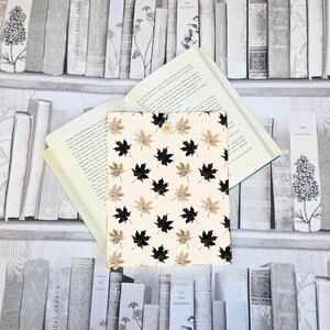 May include: A cream-coloured book cover adorned with a pattern of black and tan maple leaves. The cover is open, displaying the pages of a book. A wooden button fastens the cover. The background shows a bookshelf filled with assorted books.
