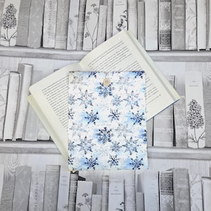 May include: A rectangular fabric book sleeve featuring a white background with a repeating pattern of blue and grey snowflakes. The sleeve has a single small wooden button closure at the top centre and is positioned over an open hardback book.