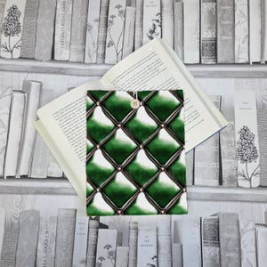 May include: A green quilted book cover with a button fastening, placed on an open book. The cover has a diamond pattern with dark green and bronze detailing. The background shows a bookshelf with various books.