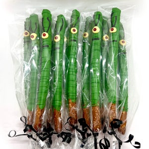 May include: A dozen green pretzel rods dipped in chocolate and decorated with a single red eye and two white eyebrows. The pretzel rods are individually wrapped in clear plastic bags and tied with black ribbon.