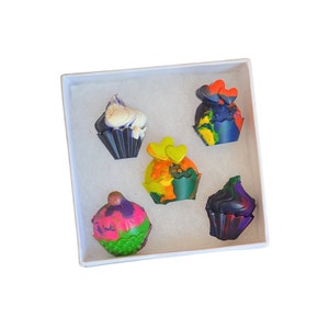 May include: Five colorful cupcake-shaped crayons in a white box. The crayons are decorated with swirls, hearts, and dots.