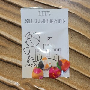 May include: A white card with the text "Let's Shell-ebrate!" and a black and white illustration of a beach ball, a sandcastle, and a shovel. There are five colorful seashells on the card.