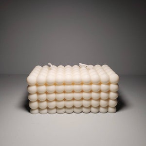 May include: A square, cream-coloured candle with a distinctive bubble pattern. The candle is made of wax and has a textured surface. Two white wicks are visible on top. A decorative item.