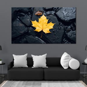 Puede incluir: Una gran impresión artística mural que presenta una hoja de arce amarilla brillante sobre rocas oscuras y mojadas. También se ve una hoja marrón más pequeña. La obra de arte se exhibe sobre un sofá negro con cojines blancos.