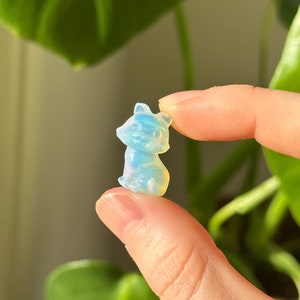 May include: A small, translucent, light blue and white fox figurine. The fox is sitting upright with its ears perked up. The figurine is held between two fingers, with a blurred green plant in the background.