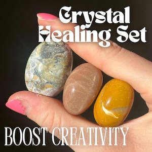 Könnte beinhalten: Ein Kristallheilungsset mit drei polierten Steinen in Grau, Pfirsich und Gelb. Der Text "Crystal Healing Set" und "BOOST CREATIVITY" wird in weißer Schrift angezeigt.