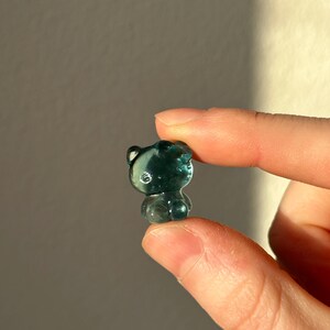 May include: A small, translucent teal-colored bear figurine. The bear has rounded features and is held between two fingers. The figurine is in focus, with a soft background. The bear is a cute, whimsical item.