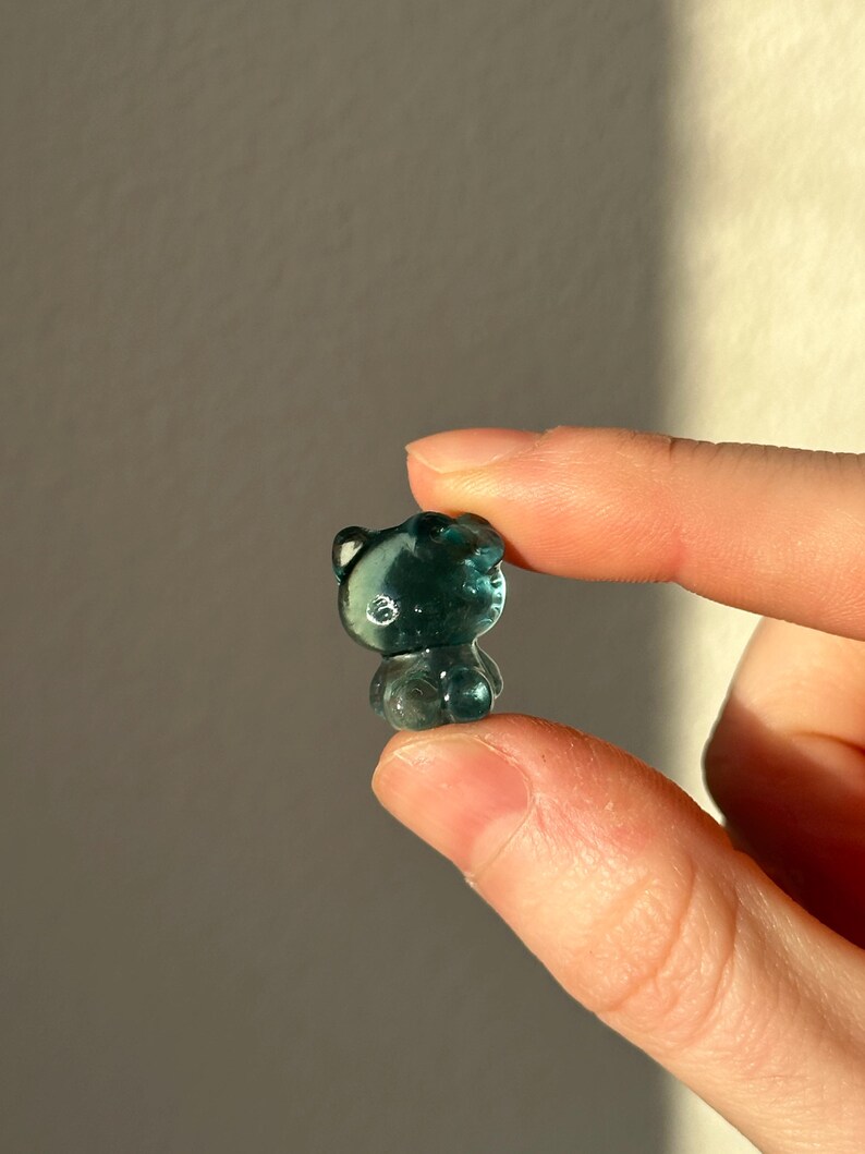 May include: A small, translucent teal-colored bear figurine. The bear has rounded features and is held between two fingers. The figurine is in focus, with a soft background. The bear is a cute, whimsical item.