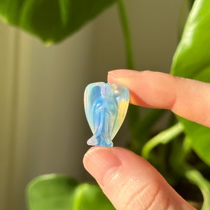 Puede incluir: Pequeña figura de ángel translúcida con un brillo iridiscente azul y amarillo. El ángel tiene las alas extendidas y se sostiene entre dos dedos. La figura está hecha de un material pulido, posiblemente cristal.