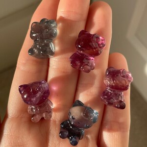 May include: Five translucent, cat-shaped figurines in shades of purple, pink, and gray. The figurines are small and appear to be made of a polished stone or crystal. The figures are held in a hand, with the background out of focus.