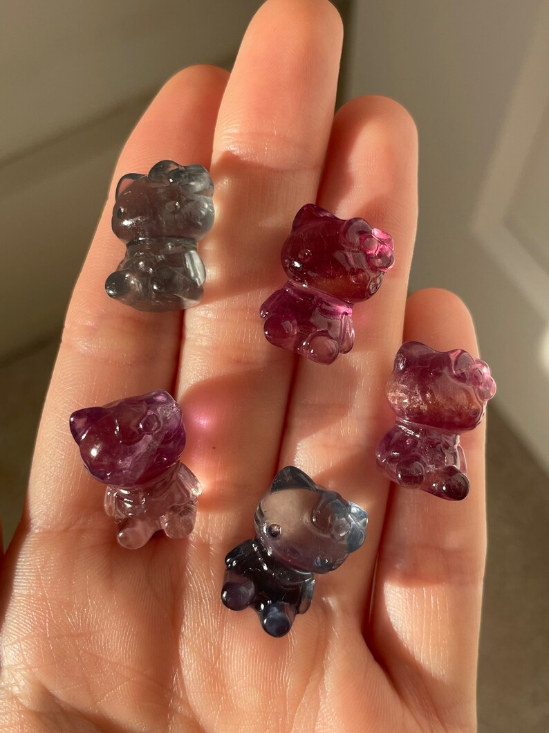 May include: Five translucent, cat-shaped figurines in shades of purple, pink, and gray. The figurines are small and appear to be made of a polished stone or crystal. The figures are held in a hand, with the background out of focus.