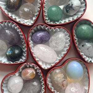 May include: A collection of polished gemstones in a variety of colours, such as green, purple, and pink, presented in red heart-shaped boxes with white paper liners. The stones have diverse textures and patterns.