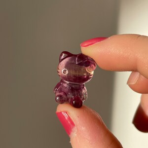 May include: A small, translucent purple figurine in the shape of a cat. The figurine has detailed features, including a bow, ears, and facial details. The cat is held between two fingers, showcasing its size and intricate design.