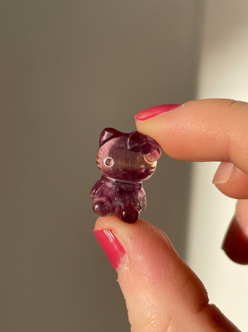May include: A small, translucent purple figurine in the shape of a cat. The figurine has detailed features, including a bow, ears, and facial details. The cat is held between two fingers, showcasing its size and intricate design.