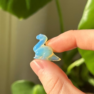 Può includere: Una piccola statuina di dinosauro opalescente. La statuina è di colore azzurro chiaro con sfumature gialle ed è tenuta tra due dita. Lo sfondo è una pianta verde sfocata.