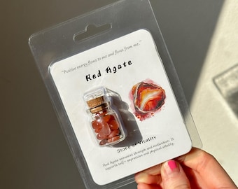 Puede incluir: Una pequeña botella de vidrio llena de piedras de ágata roja, exhibida en una tarjeta blanca. La tarjeta dice "Ágata Roja" y "Piedra de Vitalidad". El embalaje es de plástico transparente. El texto en la tarjeta dice "La energía positiva fluye hacia mí y fluye de mí."