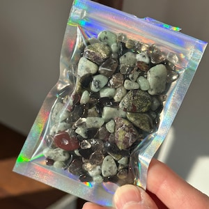 Op de afbeelding: Een doorzichtige, holografische zak gevuld met een verscheidenheid aan gepolijste edelstenen. De stenen zijn een mix van kleuren, waaronder groen, bruin, rood en helder. De zak wordt in een hand gehouden en het licht reflecteert op het holografische oppervlak.