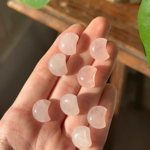 Pode incluir: Uma coleção de oito pequenas pedras polidas em forma de lua crescente de quartzo rosa. As pedras são de cor rosa suave e são seguradas na mão. O fundo inclui uma superfície de madeira e folhagem verde.