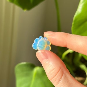 May include: A small, translucent, blue and yellow crystal shaped like a flower. The crystal is held between two fingers, with a green leafy plant in the background.