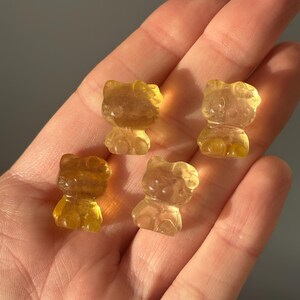 May include: Four translucent, amber-colored figurines in the shape of a cat are displayed. The figurines are held in a hand, with the background a muted gray. The cat figurines have a rounded shape with visible facial features.