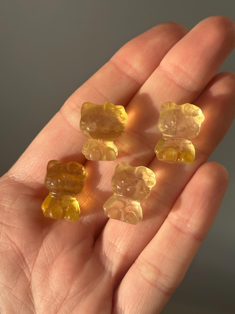 May include: Four translucent, amber-colored figurines in the shape of a cat are displayed. The figurines are held in a hand, with the background a muted gray. The cat figurines have a rounded shape with visible facial features.