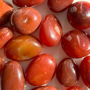 Może przedstawiać: Kolekcja wypolerowanych kamieni karneolu o różnych kształtach i rozmiarach. Kamienie mają kolory od głębokiej czerwieni do pomarańczowego, z błyszczącą, odbijającą światło powierzchnią. Niektóre kamienie mają unikalne wzory.