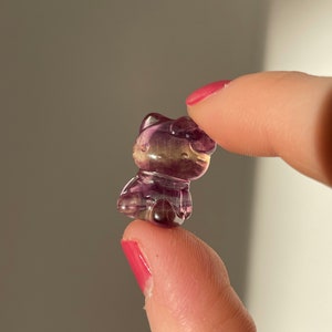 May include: A small, translucent figurine of a cat, carved from purple and yellow fluorite. The cat is held between two fingers, showcasing its detailed features and polished surface. The figurine has a cute, cartoonish design.