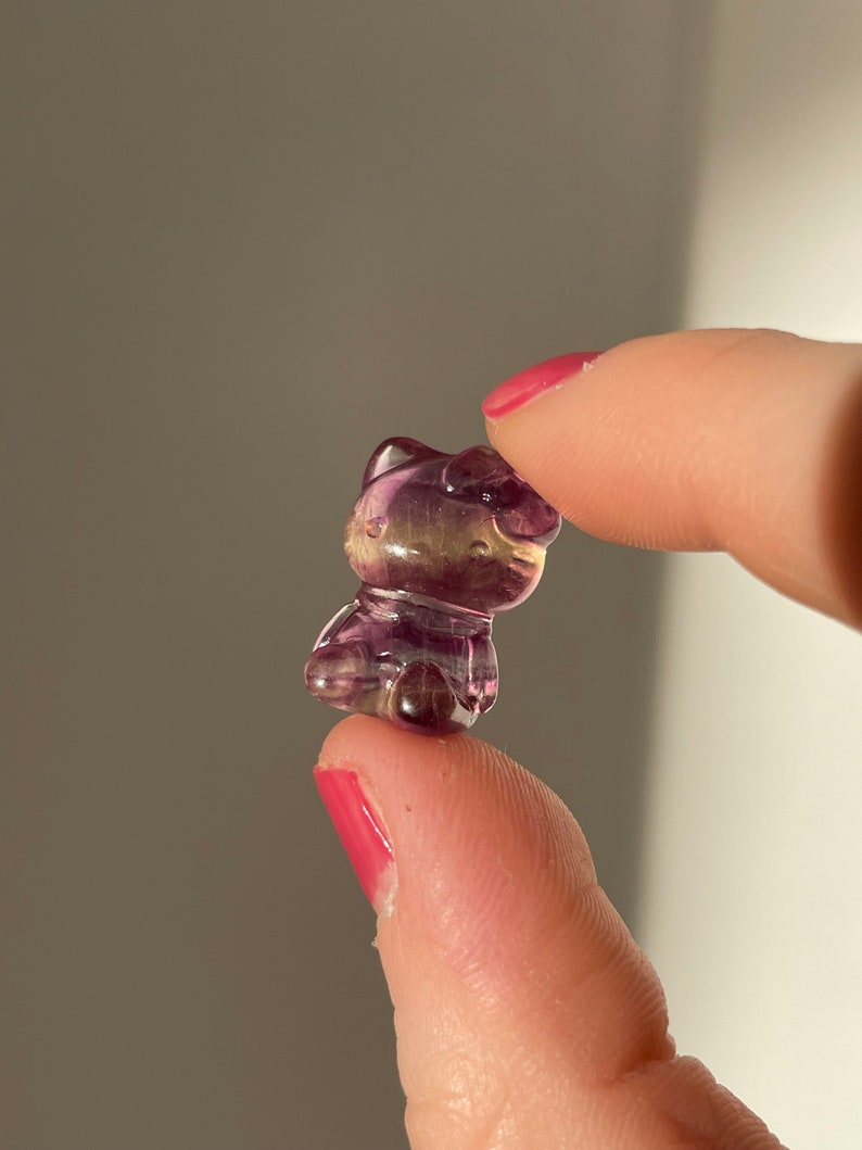 May include: A small, translucent figurine of a cat, carved from purple and yellow fluorite. The cat is held between two fingers, showcasing its detailed features and polished surface. The figurine has a cute, cartoonish design.