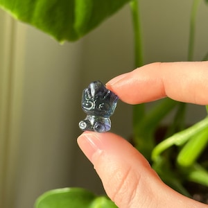 May include: A miniature, carved figurine in blue and purple hues is held between two fingers. The figurine resembles a bear or similar animal. The background is blurred, with green leaves visible.