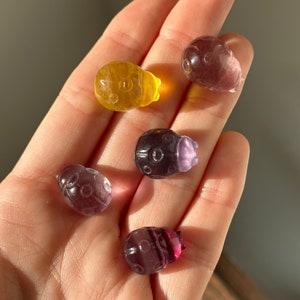 May include: A hand holds six small, carved ladybug figurines. The ladybugs are various colors, including yellow, purple, and dark red. Each has a detailed design, with circular patterns on their backs.