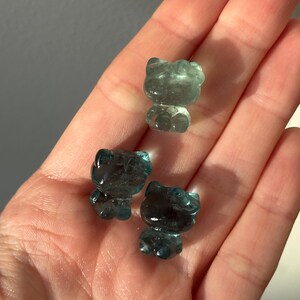May include: Three translucent blue-green cat figurines. The cat figurines are small and have a smooth, polished appearance. The figurines are displayed in a hand, with the light reflecting off their surfaces.