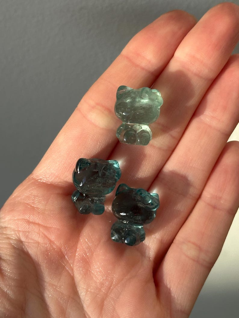 May include: Three translucent blue-green cat figurines. The cat figurines are small and have a smooth, polished appearance. The figurines are displayed in a hand, with the light reflecting off their surfaces.