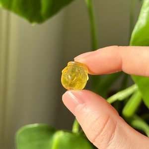 May include: A small, translucent yellow honey pot figurine. The miniature jar has a rounded shape with a lid and is held between two fingers. The background features green leaves and soft lighting.