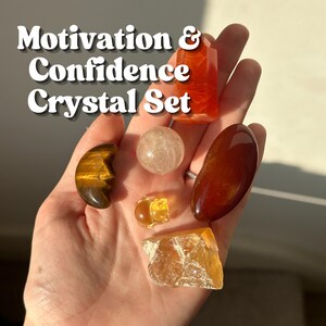 May include: A selection of crystals in different shapes and hues, including a tiger's eye crescent, a clear sphere, and a rectangular orange crystal. The text "Motivation & Confidence Crystal Set" is displayed at the top.