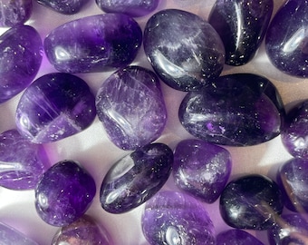 May include: A collection of polished amethyst stones in varying shades of purple. The stones are smooth, oval-shaped, and reflect light, showcasing their natural patterns and color variations. The image highlights the gemstones' rich color and texture.