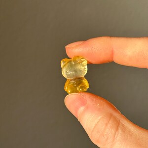 May include: A small, translucent, yellow gummy bear candy held between two fingers. The gummy bear is a classic bear shape with visible details. The background is a solid, muted gray.