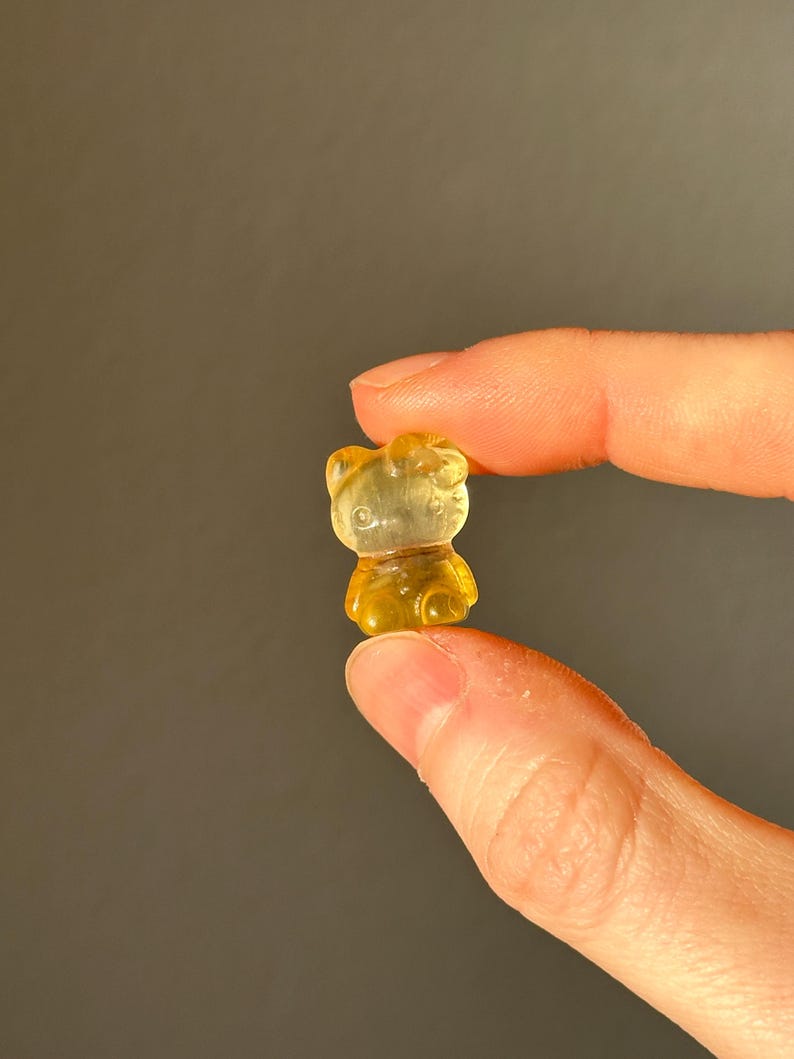 May include: A small, translucent, yellow gummy bear candy held between two fingers. The gummy bear is a classic bear shape with visible details. The background is a solid, muted gray.
