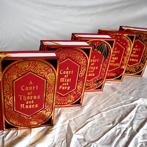 May include: A set of five red hardcover books with gold lettering and decorative designs. The titles are "A Court of Thorns and Roses", "A Court of Mist and Fury", "A Court of Wings and Ruin", "A Court of Silver Flames", and "A Court of Frost and Starlight".