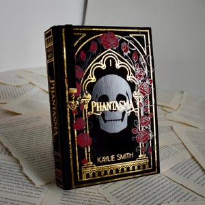 Op de afbeelding: Een zwart boek getiteld "Phantasma" met gouden details en een schedelontwerp. Rode rozen en gouden filigraan omlijsten de schedel. De naam van de auteur, Kaylie Smith, staat onderaan gedrukt. Het boek ligt op een achtergrond van gescheurde pagina's.