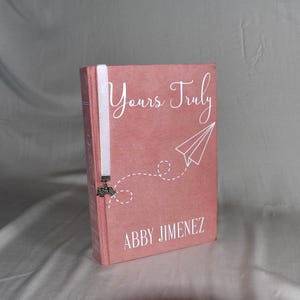 Könnte beinhalten: Ein Hardcover-Buch mit einem staubrosa Einband mit dem Titel "Yours Truly" in weißer Schrift, einer Papierflugzeug-Illustration und dem Namen des Autors, Abby Jimenez. Ein weißes Lesezeichen mit einem Anhänger ist eingelegt.